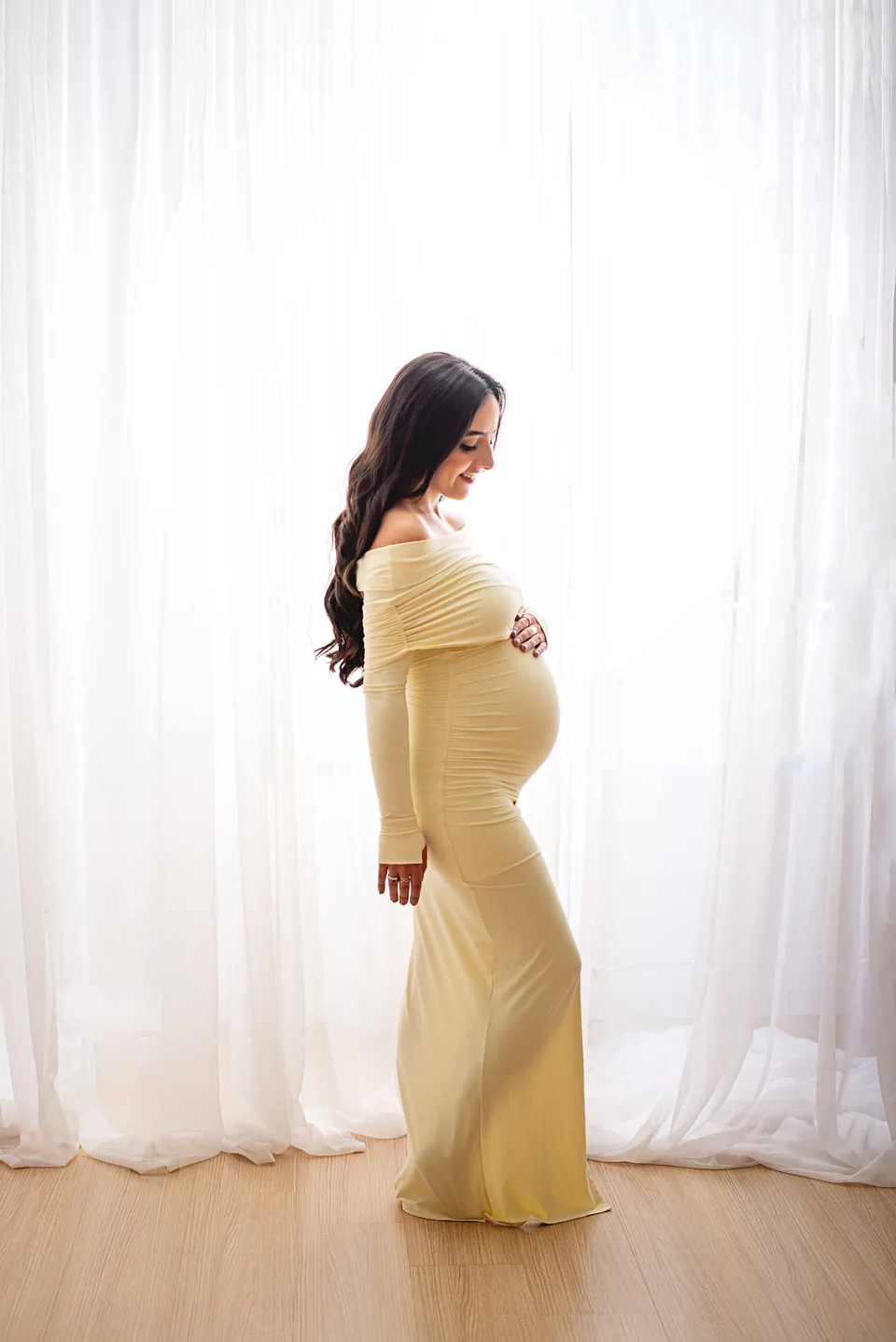 Gestante de vestido longo amarelo-claro, de perfil, olhando a barriga diante de cortinas brancas com luz natural