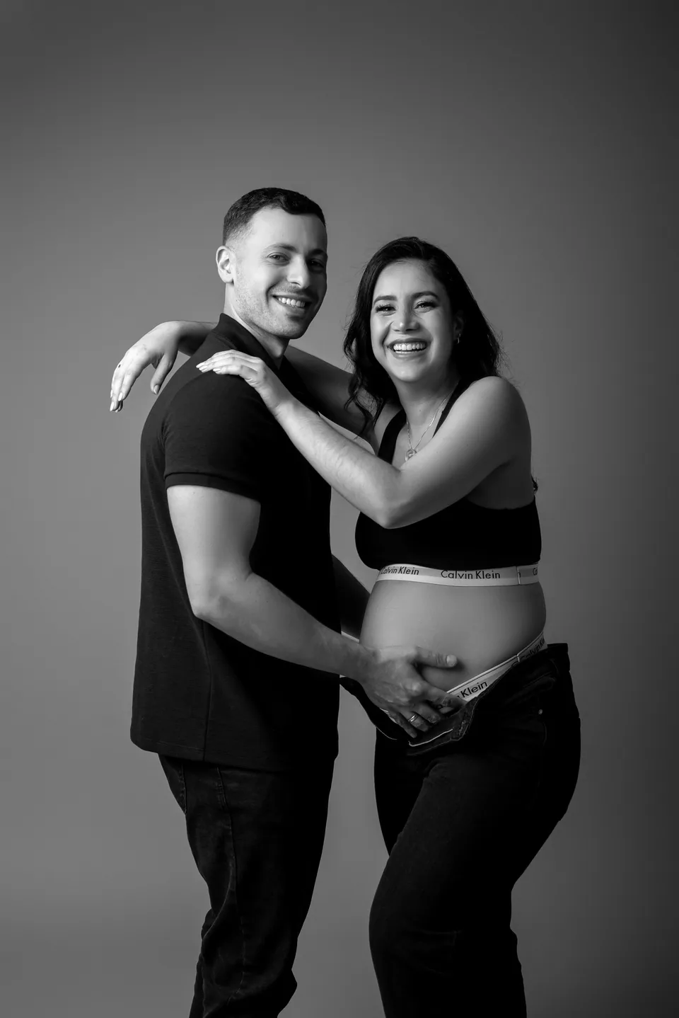 Casal abraçado sorrindo em ensaio gestante preto e branco, barriga à mostra e mão do pai sobre ela